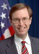 A Caucasian male posing for a professional photo wearing a gray suit with a white dress shirt and red, white, and blue striped tie. An American flag is seen in the background.