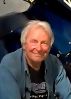 A gray haired man wearing a blue open shirt sitting at a motorcycle show in 2016