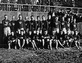 A group of 21 young men arranged in two rows—twelve standing in back and nine seated in front—in rugby uniform: dark colored sweaters emblazoned with vertically-striped shields, black belts, light colored shorts, and light colored knee-length socks.