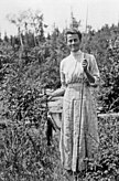 Winifred Trainor in a plaid dress, standing and facing the viewer. She is holding a string of fish in her right hand and a fly fishing rod in her left hand.