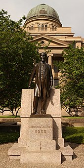 A large domed building overlooks a full-length statue of balding male with a mustache and long goatee and wearing a knee-length coat. The pedestal is engraved "Lawrence Sullivan Ross".