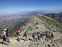 A line of students, many wearing costumes or swimwear, descends toward an alpine ridge