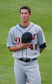 A man in a baseball uniform holds a baseball cap over his chest with his right hand.
