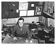 Stan Lee sits in an office, with several drawings on the background. He is sitting down in front of a table; on that table he is drawing an image.