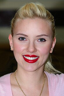 The face of a smiling woman with blonde hair pulled back into a ponytail, wearing bright red lipstick, small gold hoop earrings, a heavy gold link chain around her neck, and a pink scoop necked shirt.
