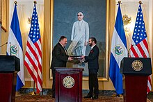 Nayib Bukele and Mike Pompeo exchanging documents in front of a painting of Óscar Romero