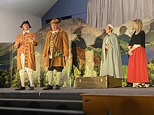 Four actors in 18th century peasant costumes stand on stage with a prop painted like a hilly landscape behind them.