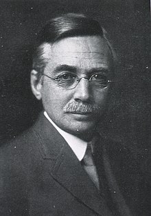 Black and white headshot of a man with a mustache and glasses wearing a suit
