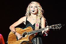 Swift playing an acoustic guitar and singing onto a mic