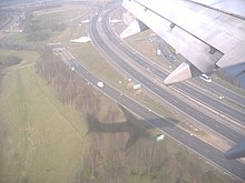 A dual carriageway and slip roads from above. Below the photographer, the well-defined shadow of an aeroplane is cast over the roads and greenery below.