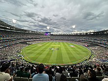 Vue panoramique de l'intérieur d'une enceinte sportive avec des tribunes remplies avant une rencontre de cricket.