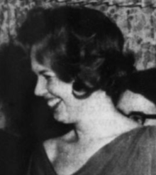 A smiling white woman in profile, with a short dark bouffant hairstyle, wearing a dark dress with a deep and wide v-neckline
