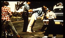 Photo of a person being arrested in the street by a policeman. The policeman is fixing the person's arms behind his back and pushing him towards a police car. One person kneeling at the side of the car is taking pictures. Two kids in the foreground are watching the scene.