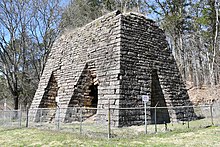 An image of a large, partially ruined, outdoor stone furnace with two bays for smelting iron surrounded by a chain link fence.