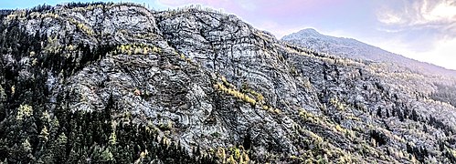 Photographie d'une falaise où on voit de nombreux plis de géologie gris clairs, qui se déversent de chaque côté, légèrement forestiers et en barres rocheuses semblables à des strates horizontales et déformées.