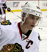Jonathan Toews in a white away Blackhawks jersey and wearing a white helmet with a clear shield covering his eyes.