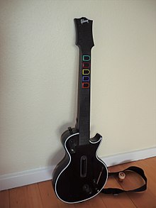 A black guitar controller sits on a white piece of paper and a wooden floor.