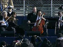 Four male musicians playing a variety of instruments outdoors in front of a chain-link fence
