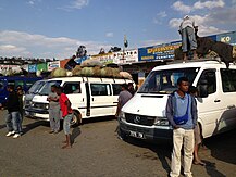 Four vans are parked in parallel in front of a series of storefronts. Some have goods on top in woven containers. Men are standing on top of two of them, loading or unloading them. Other men congregate nearby.