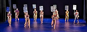 Dancers in brown facing the audience, holding handwritten black-and-white signs saying "no justice," "no peace," and "can you hear me?"