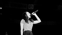 A black-and-white image. Against a black background, a young woman in side profile holds a microphone high and belts into it.