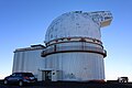 The telescope building is pictured against a blue sky. The left side of the building is rectangular, while the right side is cylindrical and topped with the dome. The dome has a protrusion extending rightward. In the middle of this side of the building is the entrance. A black SUV is parked to the left of the entrance, and a white Jeep is parked on the horizon to the right of the building.