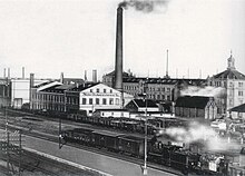 Černobílá fotografie s šikmo ubíhající silnicí v popředí a s budovou nádraží, za níž je komplex budov továrny s kouřícím komínem