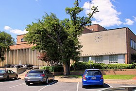 Vue d'un bâtiment avec une horloge sur la façade. Devant le bâtiment se trouve un arbre ainsi qu'un parking et des automobiles.