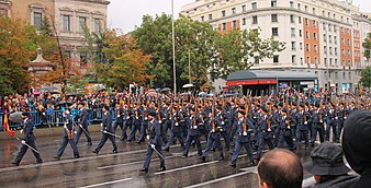 Several dozen male soldiers in formal steel blue uniforms carrying wooden rifles march down a wide street while a crowd looks on
