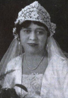 A young Black woman wearing a bridal ensemble, including a lace crown-shaped cap, a veil, a white jacket, and a lace dress