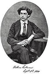 Sullivan seated with one leg crossed over another, age 16, in his Royal Academy of Music uniform, showing his thick, curly hair. Black and white.