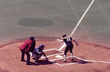 A view of home plate from the first base side seats. A left-handed batter with the number 48 on his black uniform is in the batter's box looking down at home plate with one hand on the bat which is resting on his shoulder. The catcher in a white uniform has just caught a ball close to the ground. The umpire, wearing blue pants and a red jacket, is leaning over the catcher.