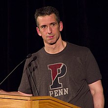 A 47-year-old white man is standing behing a lectern, looking to the camera's right; he has dark hair, and is wearing a grey t-shirt that says "PENN"