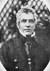 Photographie noir et blanc, en extérieur sur fond de treille, en buste d'un homme blanc aux cheveux clairs et habit sombre