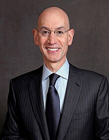 A thin, bald man in a dark pinstripe suit, wearing glasses, striped shirt and dark blue tie against a gray background