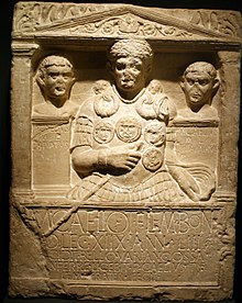 A stone carving, the border resembling a Greco-Roman building, displaying three busts, slightly damaged, the centremost being that of Marcus Caelius, wearing armour, a cape and holding a staff of office in his right hand. Below the carving is a Latin inscription.