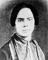 Photographie noir et blanc en portrait d'une femme blanche avec des cheveux sombres en bandeaux, toilette boutonnée jusqu'au cou