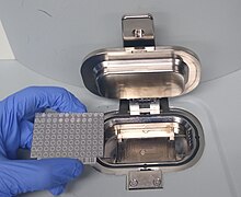 A gloved hand holds a metal plate onto which microbial samples have been placed, ready to load it into the sampling area of the MALDI-TOF instrument