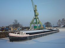 Barevná fotografie zachycují zmrzlou řeku s lodí a nákladním jeřábem