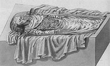An open tomb seen from the side in a 45-degree angle from the ground. The corpse, with his head to the left, is dressed in fine funeral attire, wears a coronet and holds a sceptre in each hand.