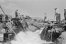 Four men dressed in long-sleeved shirts, long pants, and hats are perched on platforms on both sides of a rushing stream. Three of the men are standing, and one is seated. Each man holds one end of a long pole with a net, dipped in the water, attached to the other end. Several people without poles are watching or waiting nearby.