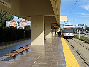 A light rail train at a station with a blocky concrete canopy