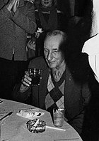 A black-and-white photograph of an older man sitting at a table and drinking a glass, surrounded by people