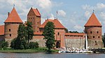 Red brick castle in the background. Lake in front. A wooden bridge with tourists. Sailboats on the lake.