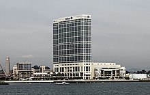 Photographie en couleurs représentant un bâtiment blanc aux nombreux étages, à la façade vitrée, sur le front de mer.