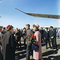 Kennedy and the First Lady, dresseing in a pink outfit and holding a bouquet of flowers, depart from Air Force One and greet welcomers.