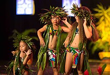 Trois enfants dansent sur scène, vêtus de costumes de tissu et de feuilles, avec des couronnes végétales sur la tête
