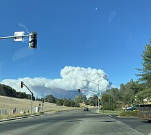 A large gray and white plume of smoke rises in the distance beyond a line of trees, seen from behind the windshield of a vehicle at an intersection