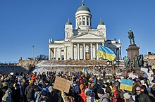 A crowd of people in winter coats march past a white domed church above a set of snowy stairs, some carrying signs and blue and yellow Ukrainian flags.