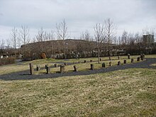 A grassy area with stones arranged in an oval shape demarcating a burial area. In the distance can be seen trees and a hill.